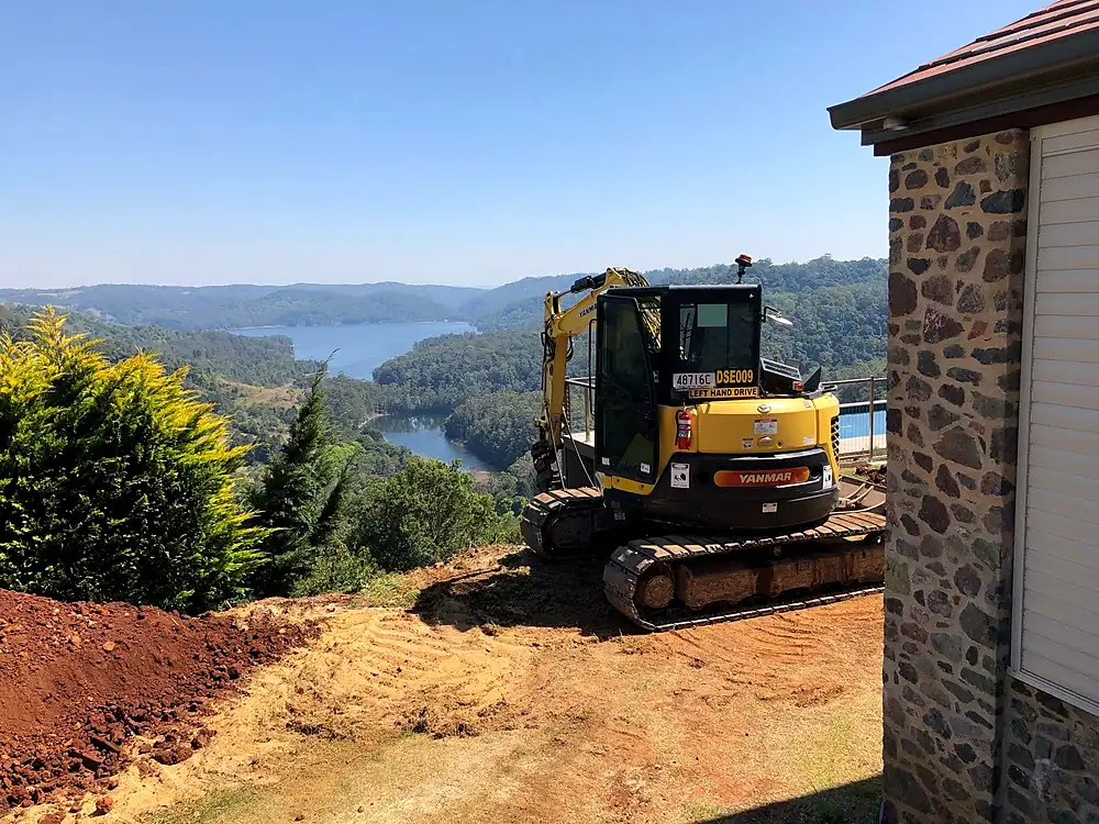 Excavator on a cut hillside pad beside a stone house above a lake