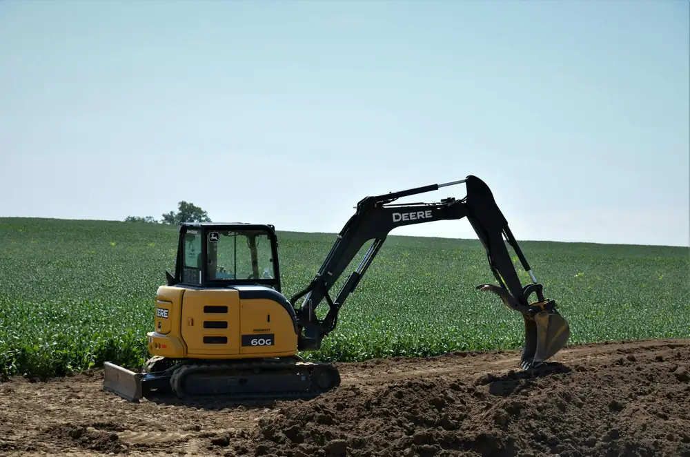 Compact yellow excavator working on a cleared dirt patch beside a green field